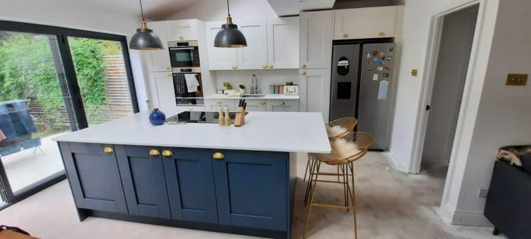 Modern kitchen interior featuring a stylish kitchen island with a white countertop, blue cabinetry, and brass handles. The kitchen includes built-in appliances, pendant lighting, and bar stools. An Arch KBB team member is present, showcasing the functional yet aesthetic design of the space.