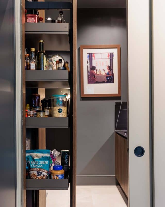 Interior view of a modern pantry showcasing neatly arranged food items and condiments on shelves, with a framed photograph on the wall. An Arch KBB team member is present, demonstrating the functionality of the space.