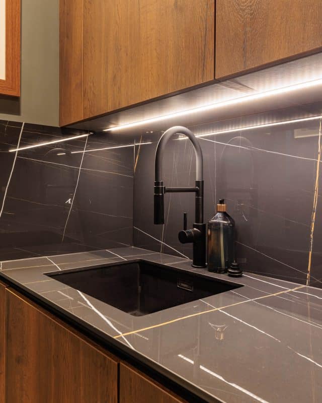 Modern kitchen sink with a sleek black tap and stylish accessories, featuring a dark stone countertop with gold veining and ambient lighting above. An Arch KBB team member is visible in the background, contributing to the design.