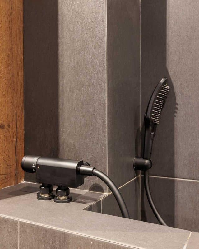 Close-up of a modern bathroom feature showcasing a sleek black showerhead and a comb attachment, set against stylish grey and wooden wall finishes. An Arch KBB team member is present, demonstrating the innovative design and functionality of the installation.