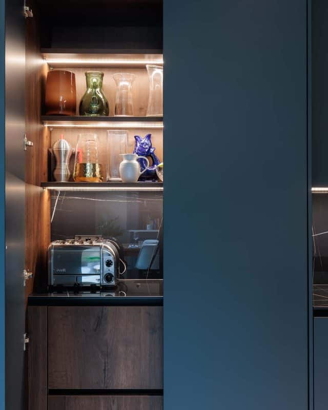 Arch KBB team member showcases a modern kitchen cabinet featuring a stylish toaster, an array of decorative glassware, and vibrant ceramics, all illuminated by sleek integrated lighting.