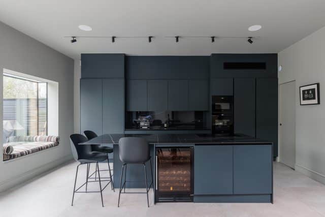 Modern kitchen design featuring dark cabinetry, sleek appliances, and an island with bar stools. The space is well-lit with track lighting and includes a window seat for added comfort. An Arch KBB team member is present, showcasing the stylish layout.
