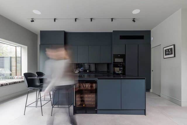 Modern kitchen featuring sleek dark cabinetry, integrated appliances, and an elegant design. An Arch KBB team member is seen in motion, adding a dynamic element to the contemporary space. The room is well-lit with track lighting and includes minimalist bar stools, enhancing the kitchen's stylish atmosphere.