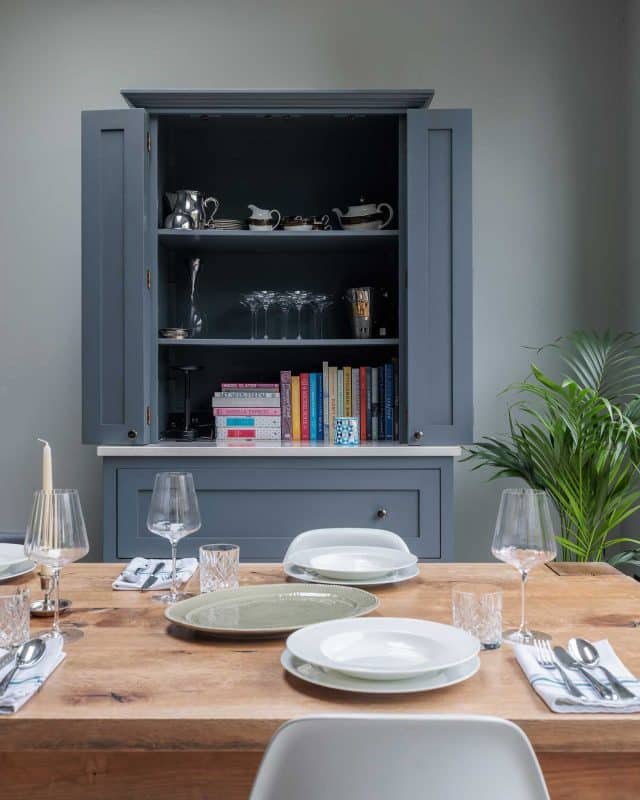 A well-set dining table with elegant glassware and plates, featuring a stylish grey cupboard in the background displaying glassware and books, accompanied by a decorative plant on the side. An Arch KBB team member is present, contributing to the inviting atmosphere.