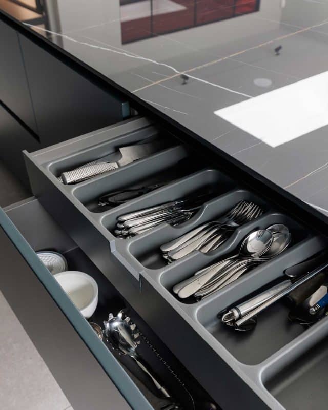 A neatly organised kitchen drawer featuring a variety of utensils, including knives, forks, spoons, and a grater, all arranged in a grey cutlery tray. The drawer is part of a modern kitchen design showcased by an Arch KBB team member.