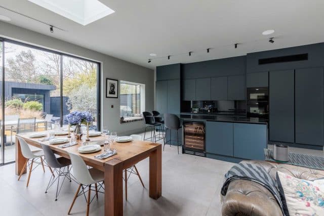 Modern kitchen and dining area featuring sleek grey cabinetry, a wooden dining table set for a meal, and contemporary seating. Large windows provide natural light and a view of an outdoor space with greenery. An Arch KBB team member is present, adding to the inviting atmosphere.