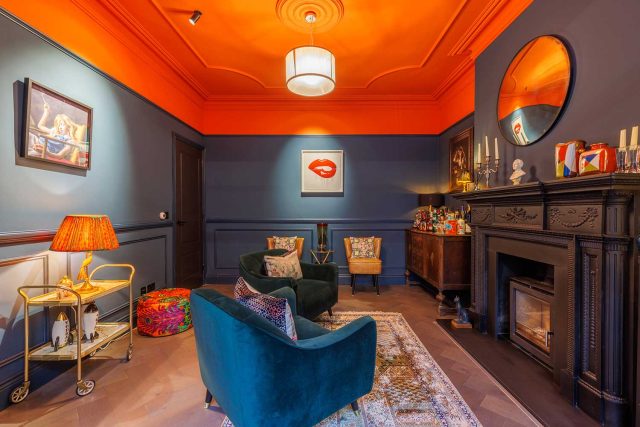 Stylish interior of a living room featuring bold orange and dark blue walls, elegant furnishings, and decorative accents. The space includes a fireplace, a round mirror, and artwork, creating a vibrant and inviting atmosphere. An Arch KBB team member is present, contributing to the room's aesthetic appeal.