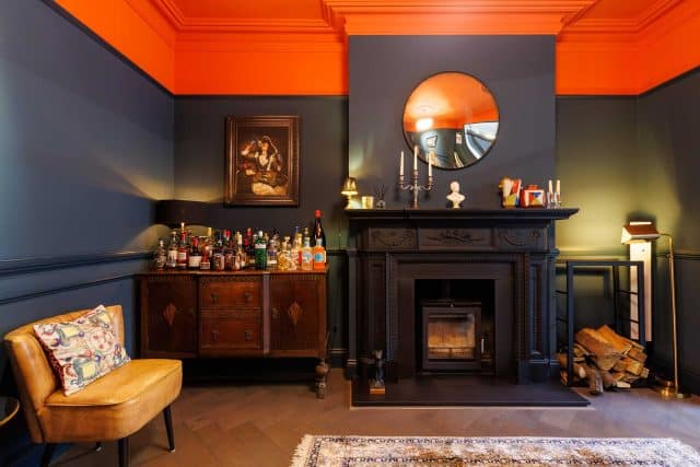 Interior of a stylish living room featuring a dark blue and orange colour scheme. An ornate black fireplace serves as a focal point, adorned with candles and decorative items. A wooden cabinet stocked with an array of spirits sits alongside a comfortable chair and a patterned rug, creating an inviting atmosphere. An Arch KBB team member is present in the space.