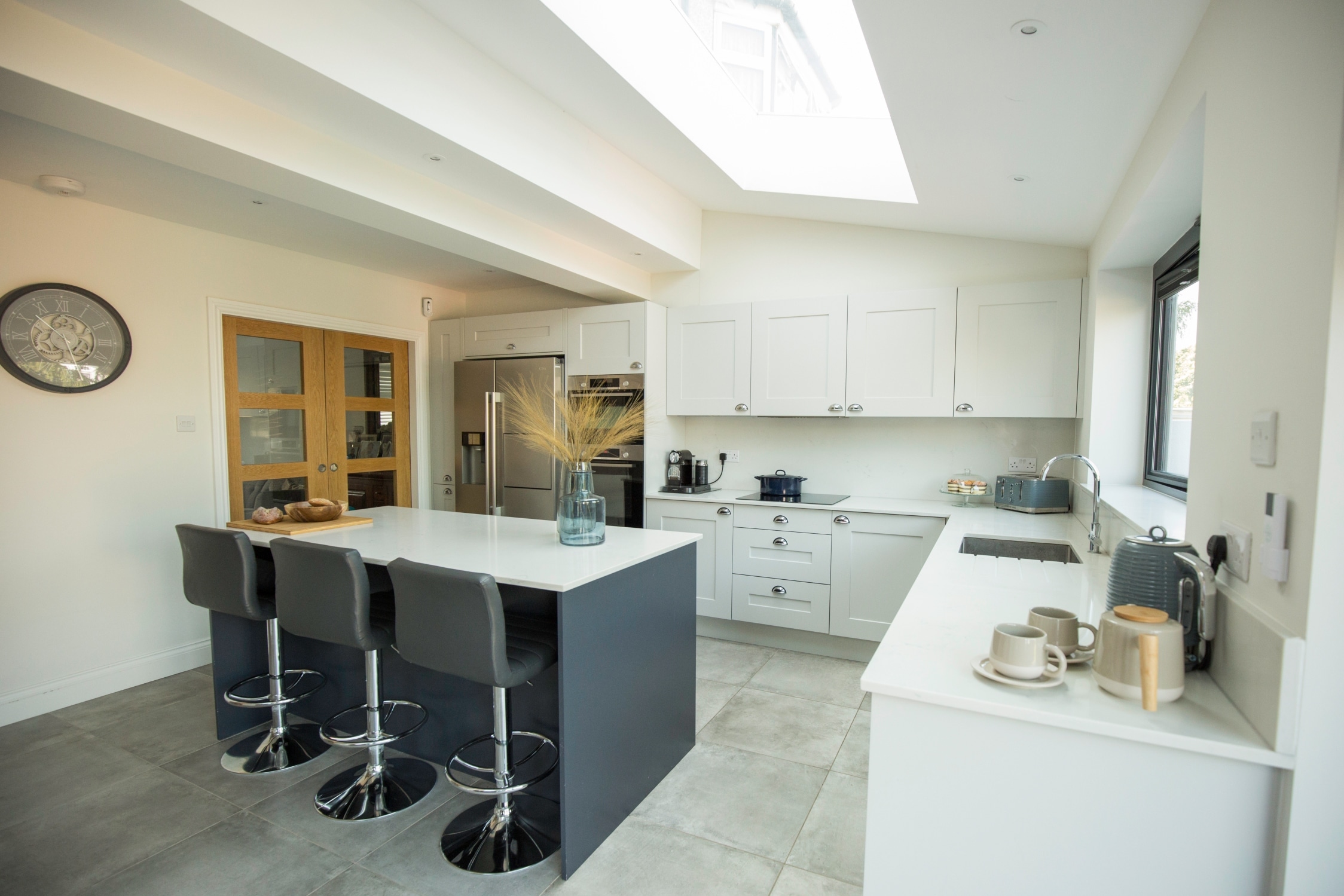 Modern kitchen featuring a spacious layout with a central island and stylish bar stools. The design includes sleek cabinetry, stainless steel appliances, and large windows allowing natural light to illuminate the space. An Arch KBB team member is present, enhancing the kitchen's inviting atmosphere.