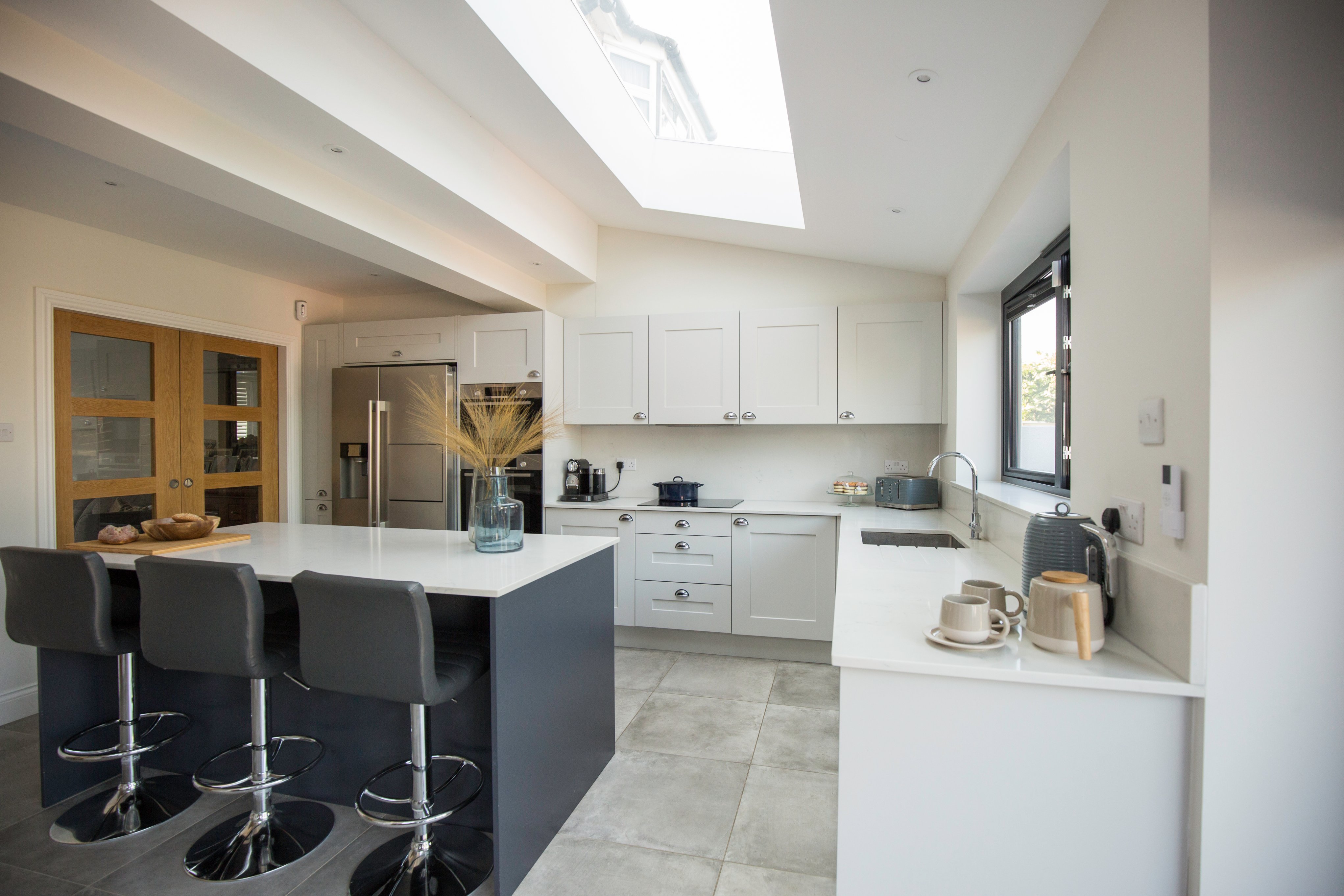 Modern kitchen design featuring sleek cabinetry, a central island with seating, and ample natural light from a skylight. The space includes stainless steel appliances, minimalist decor, and a functional layout, ideal for cooking and entertaining. An Arch KBB team member is present, showcasing the kitchen’s inviting atmosphere.