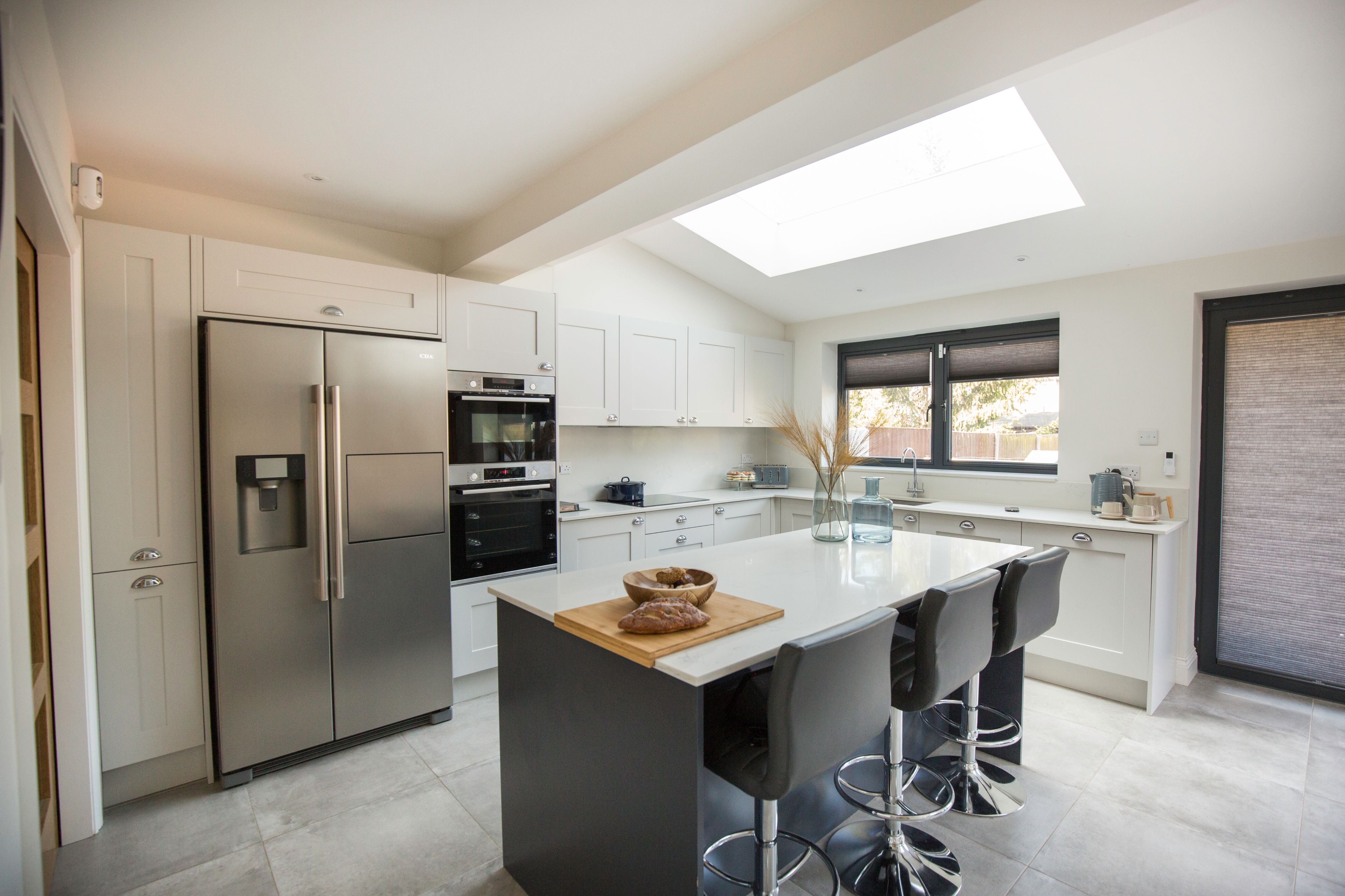 Modern kitchen featuring sleek cabinetry, stainless steel appliances, and a spacious island with bar stools. Natural light floods the room through a skylight, enhancing the contemporary design. An Arch KBB team member can be seen, adding a personal touch to the space.