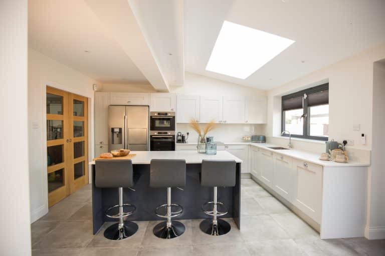 Modern kitchen design featuring a spacious layout with white cabinetry and a central island. The island is equipped with stylish bar stools, complemented by contemporary appliances including a refrigerator and oven. Natural light floods the room through a skylight, enhancing the bright and airy atmosphere. An Arch KBB team member is present, showcasing the kitchen's functionality and aesthetic appeal.