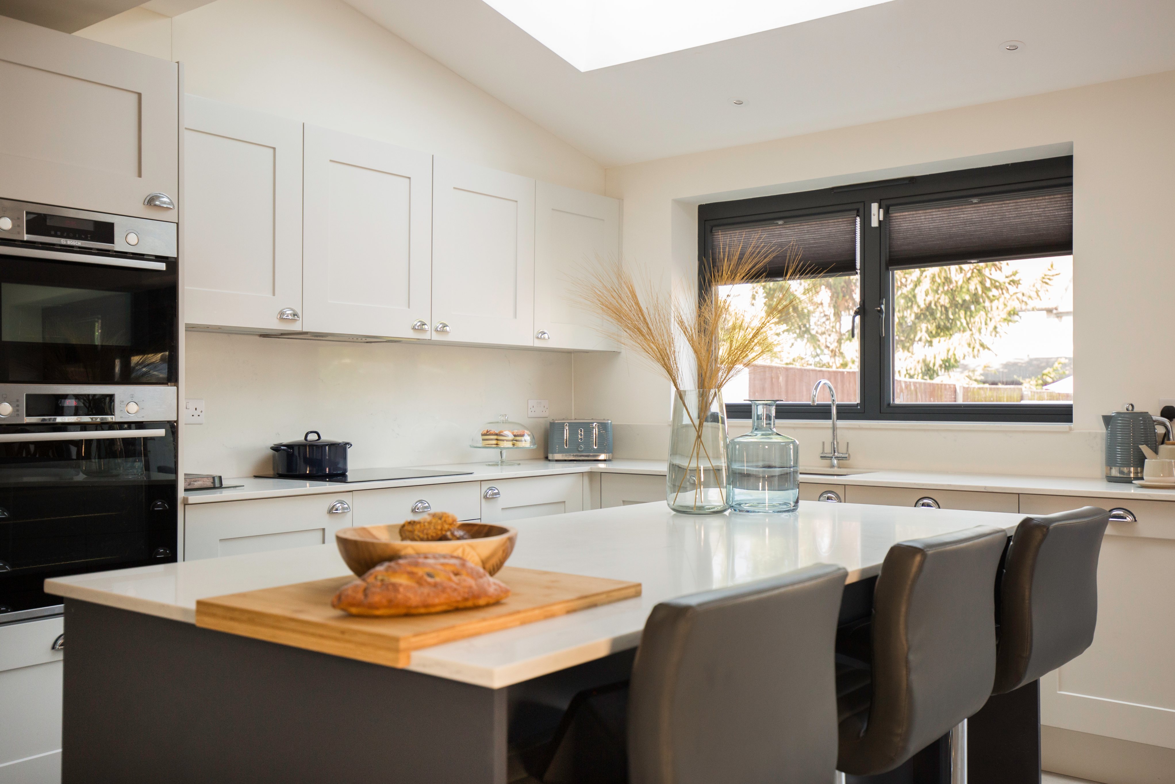 Modern kitchen interior featuring sleek cabinetry, a central island with a wooden chopping board displaying bread, and decorative elements like dried grass in a vase. An Arch KBB team member is present, enhancing the inviting atmosphere of the space.