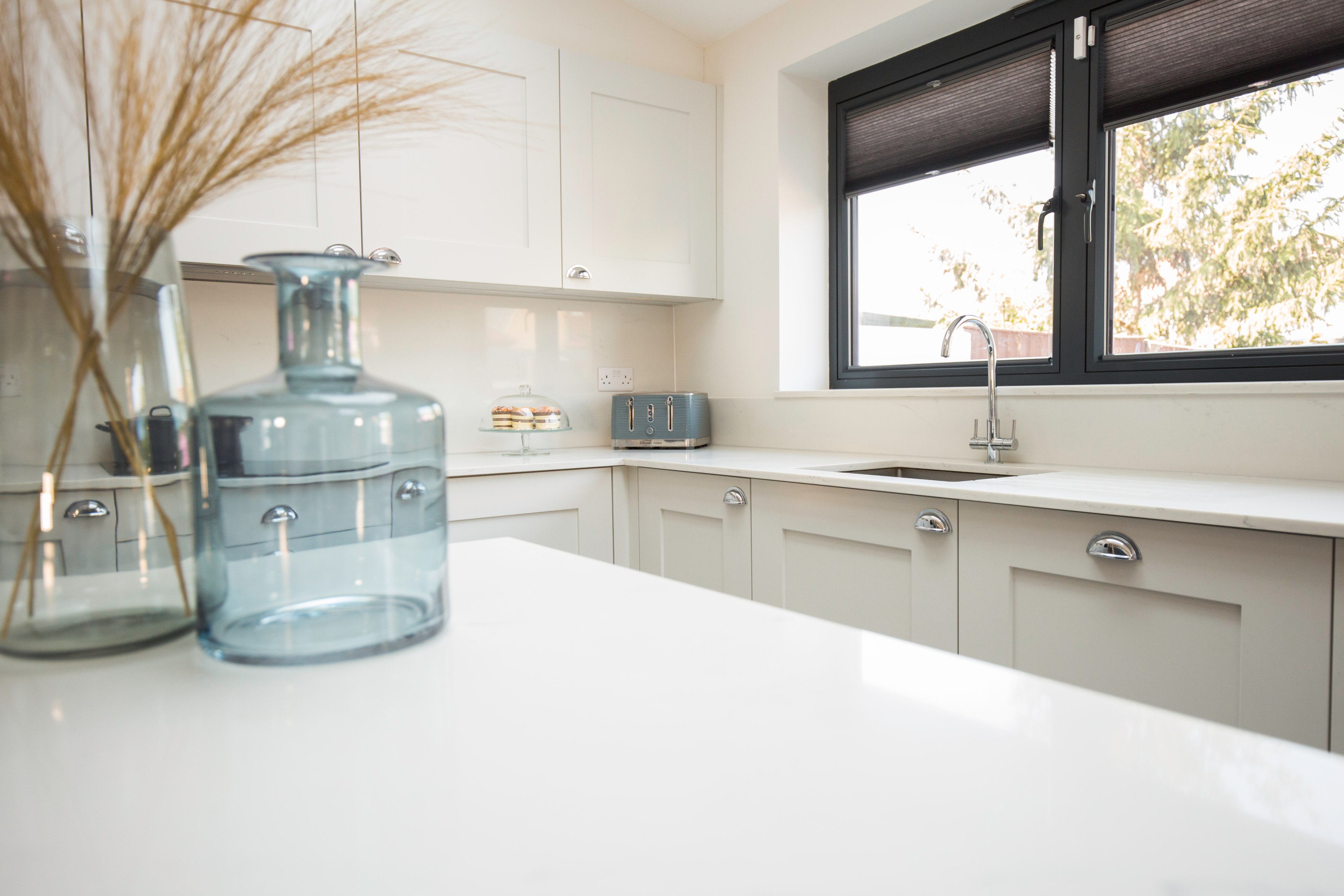 Modern kitchen interior featuring sleek cabinetry, a minimalist design, and natural light streaming through the window. A decorative glass vase and a cake stand add a touch of elegance to the space, with an Arch KBB team member present to enhance the kitchen's inviting atmosphere.