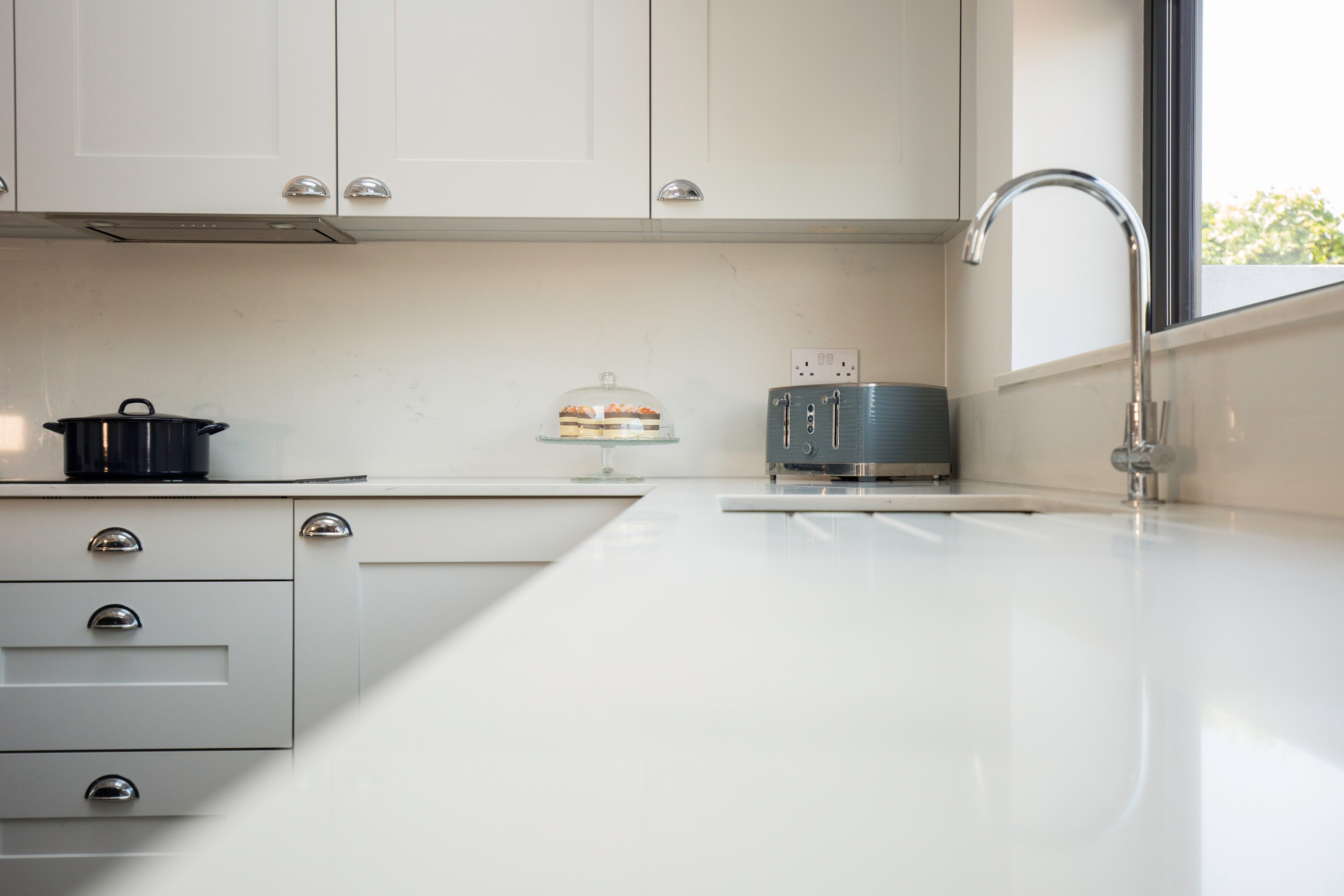Modern kitchen design featuring sleek white cabinetry, a polished countertop, a kettle, and a toaster. A glass cake stand with pastries is visible, enhancing the inviting atmosphere. An Arch KBB team member is present, contributing to the scene.