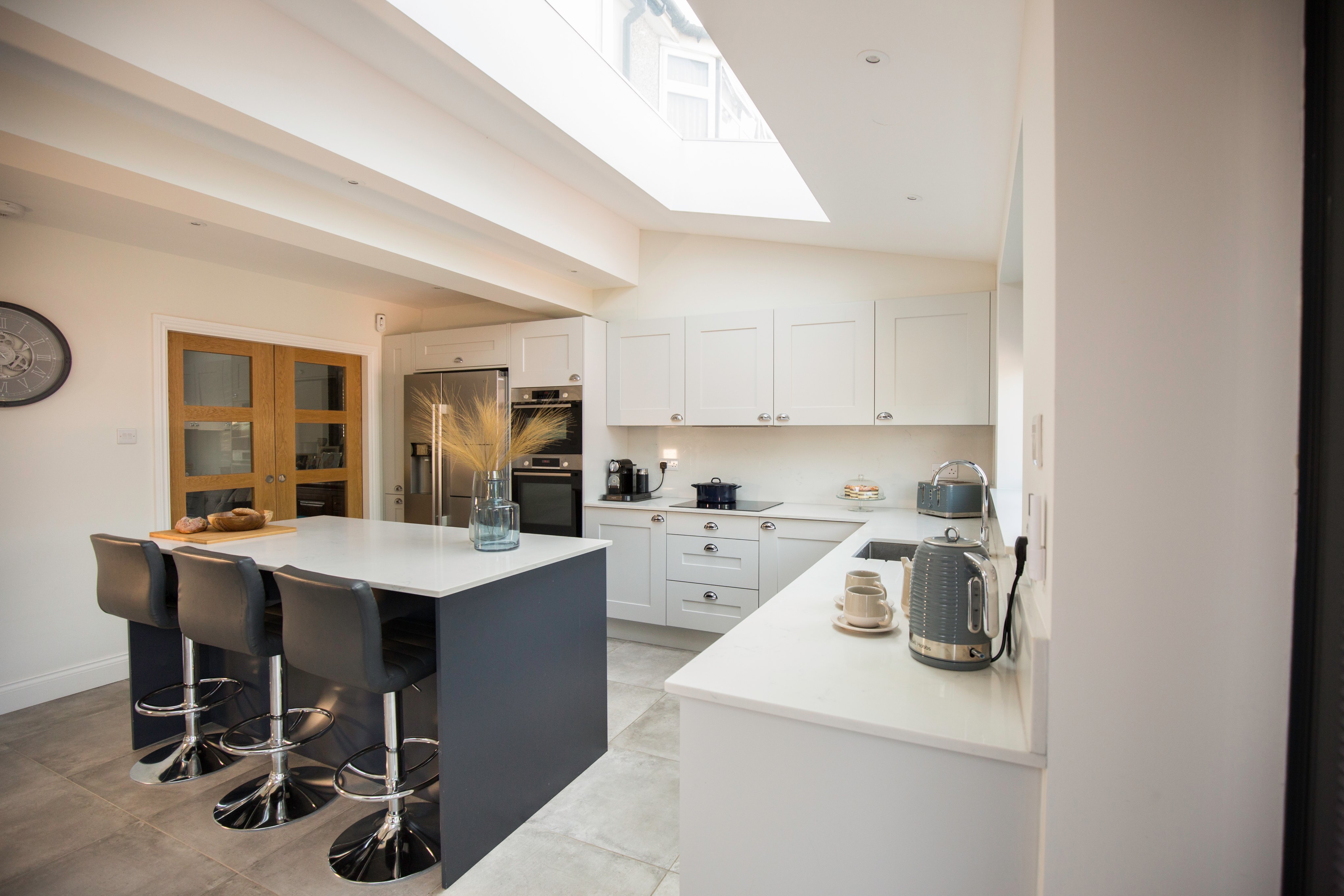 Modern kitchen design featuring a spacious layout with a central island and bar stools, sleek cabinetry, and integrated appliances. Natural light floods the space through a skylight, enhancing the contemporary aesthetic. An Arch KBB team member is present, showcasing the kitchen's functionality and style.