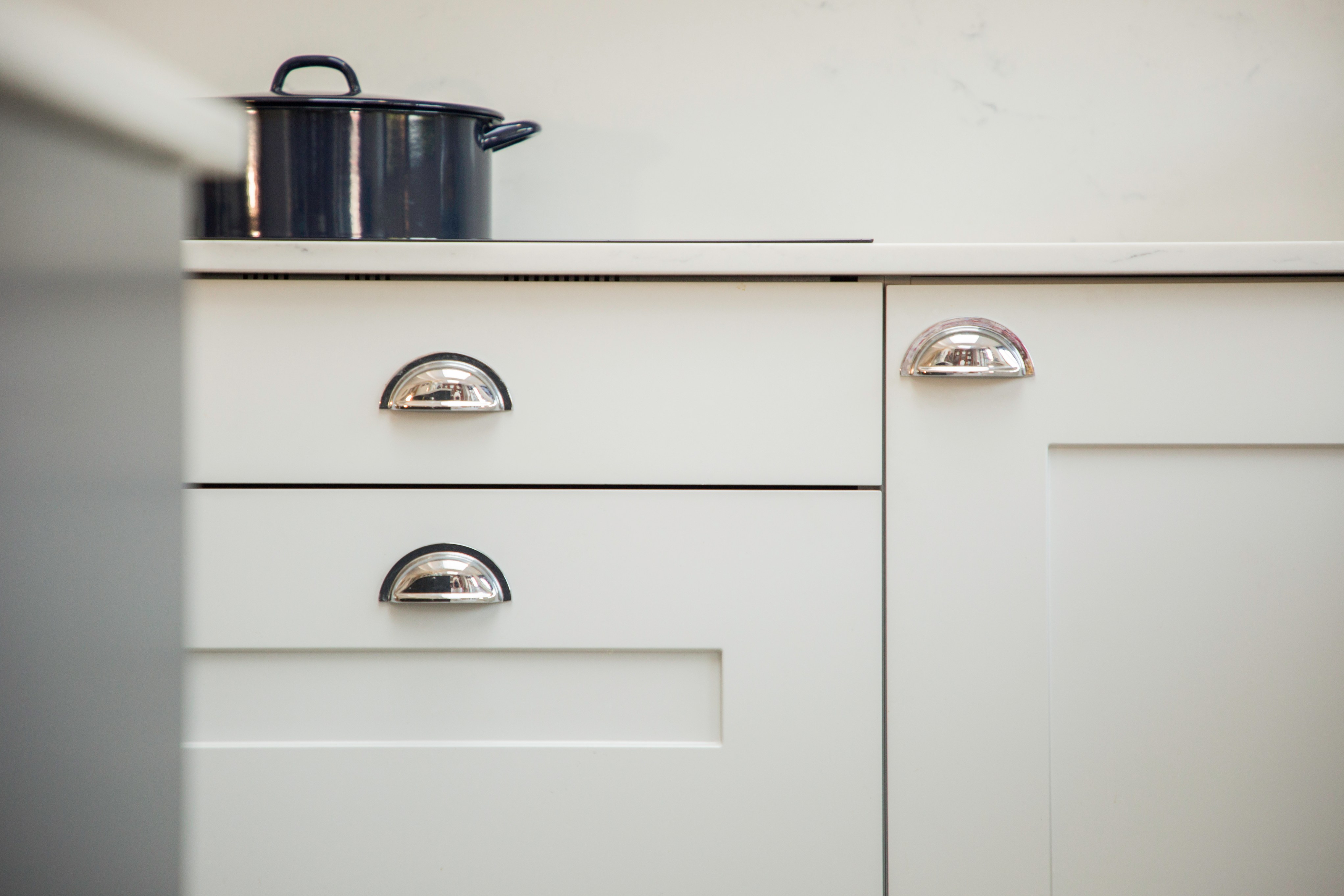 Close-up view of a modern kitchen featuring white cabinetry and shiny chrome handles, with a black cooking pot on the countertop, showcasing a sleek and contemporary design by Arch KBB team member.