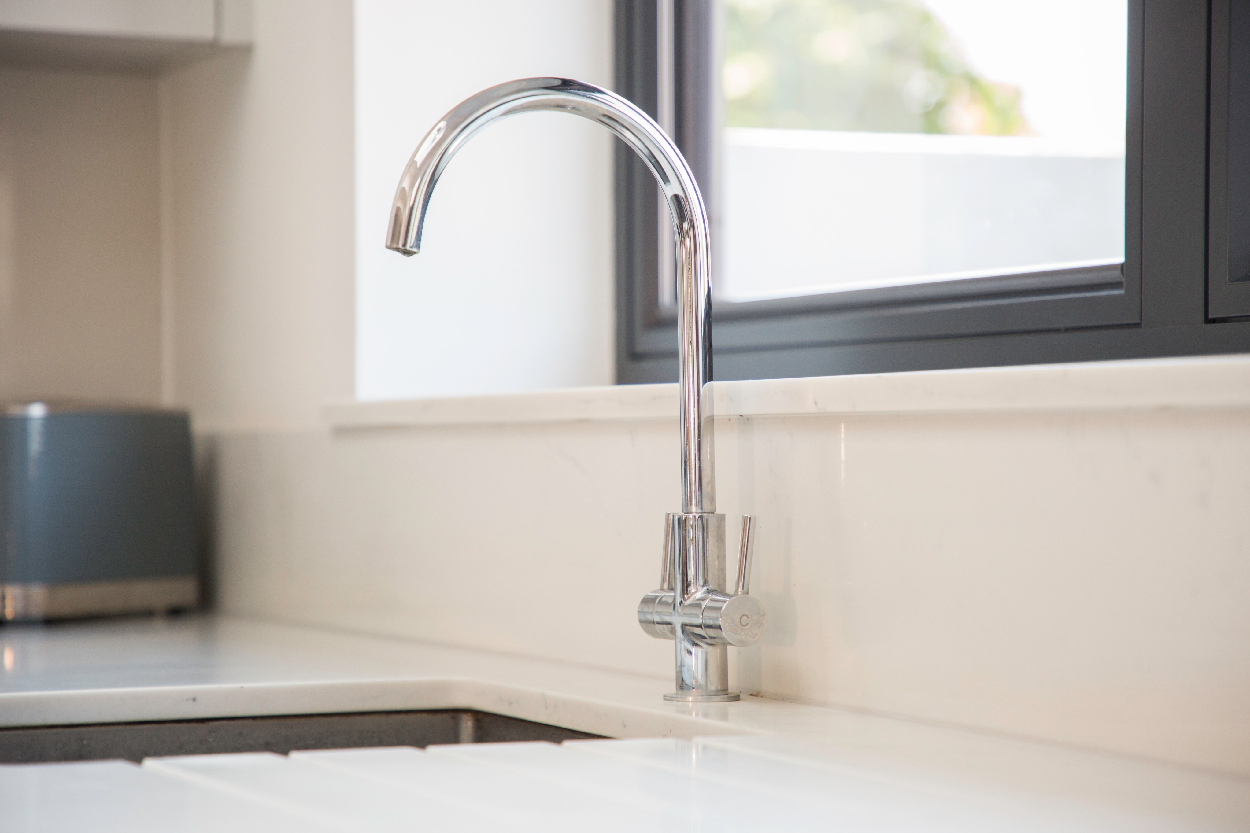 A sleek chrome kitchen tap positioned on a modern countertop, featuring a minimalist design and set against a backdrop of a window, allowing natural light to illuminate the space. Ideal for contemporary kitchen aesthetics, showcasing quality and functionality.