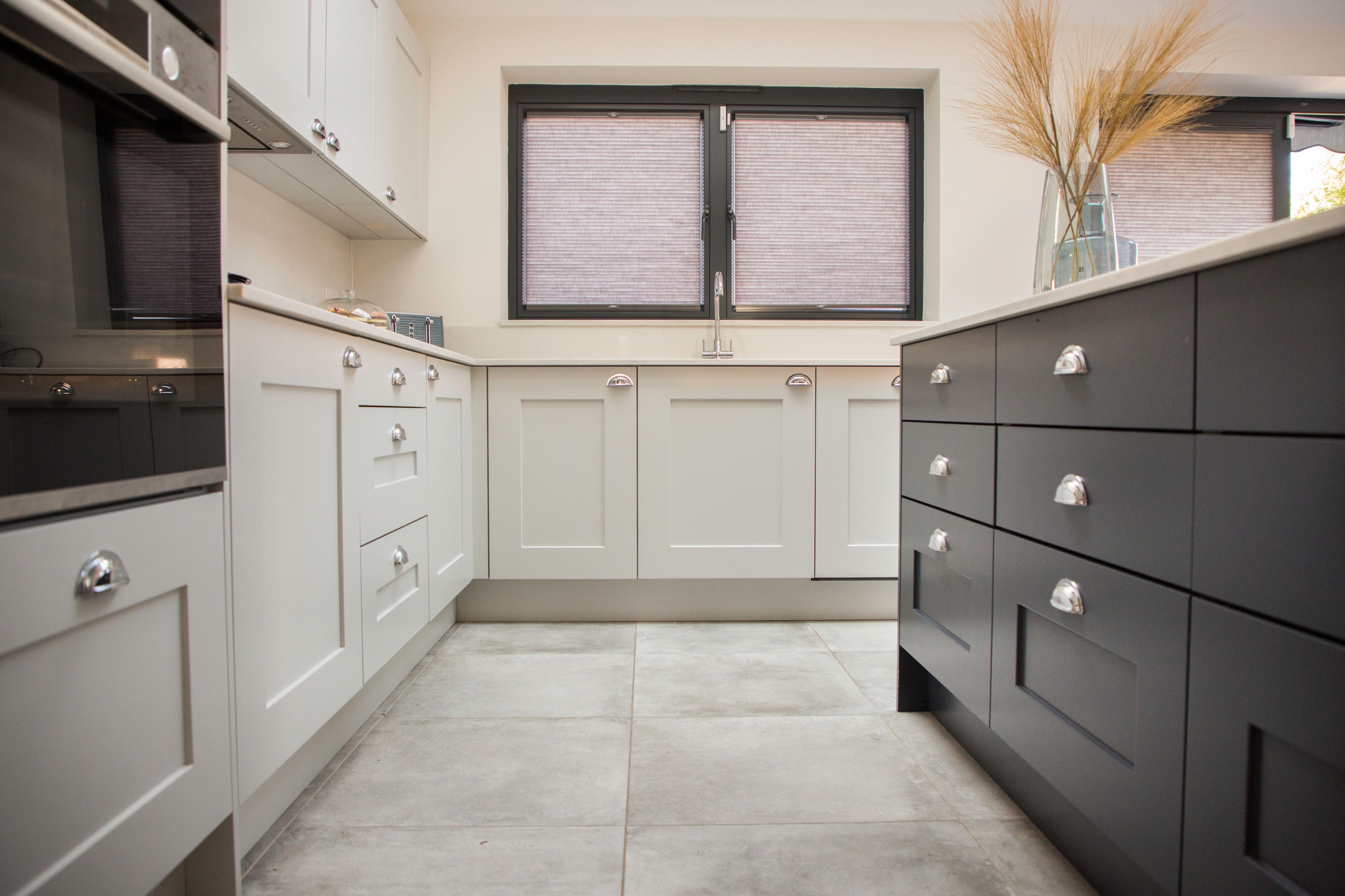 Modern kitchen design featuring a combination of grey and black cabinetry, sleek handles, and a spacious layout. Natural light flows through the large window, illuminating the stylish tiles on the floor. An Arch KBB team member is present, showcasing the kitchen's functionality and aesthetic appeal.