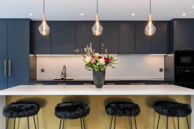 Modern kitchen featuring a sleek design with dark cabinetry, a marble backsplash, and stylish pendant lighting. A central island with a white countertop is adorned with a floral arrangement in a black vase, complemented by elegant bar stools with fur detailing. An Arch KBB team member is present, showcasing a contemporary kitchen aesthetic.