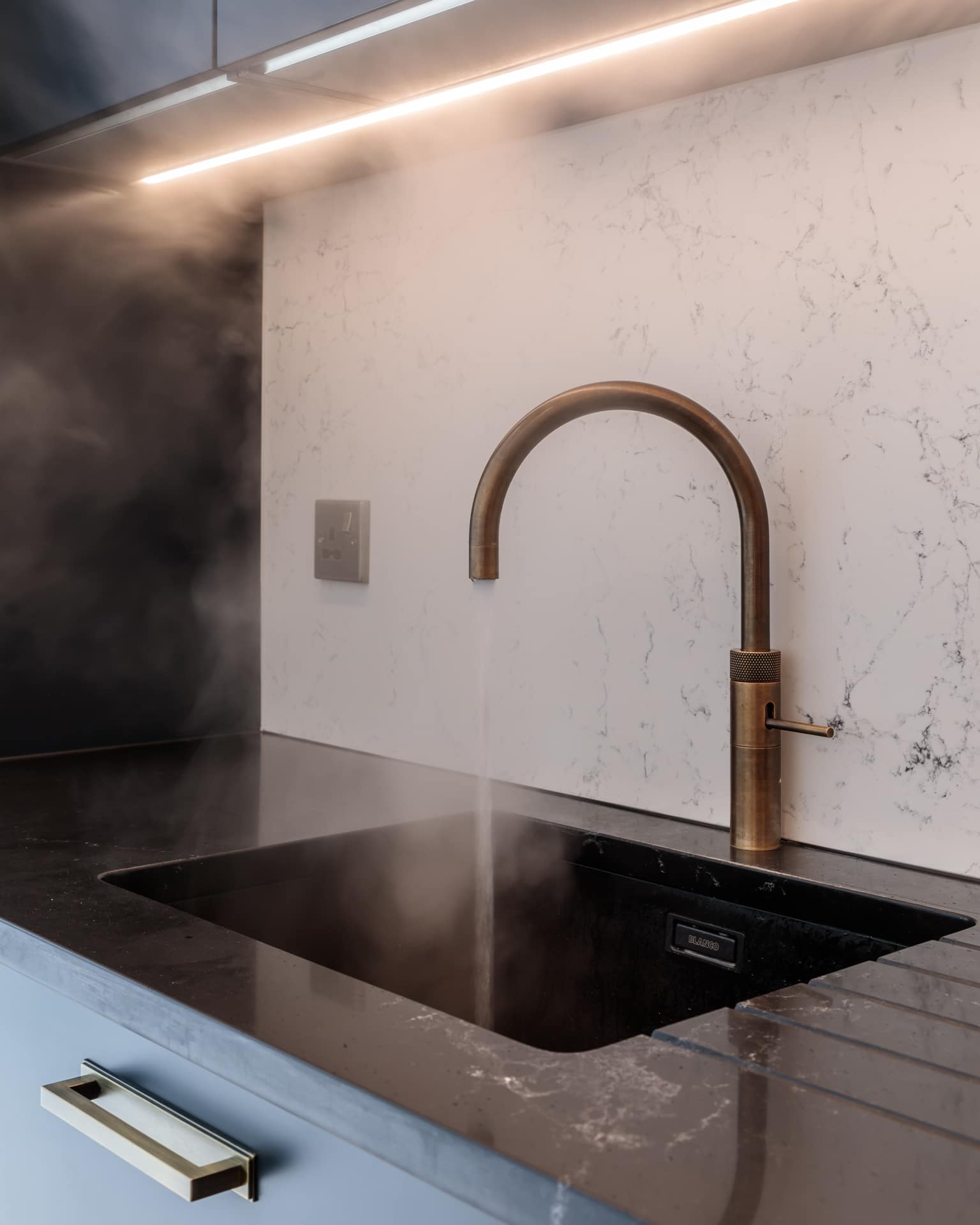 Modern kitchen featuring a stylish bronze tap with steam rising from a black sink, set against a marble backsplash. An Arch KBB team member is present, showcasing contemporary kitchen design elements.