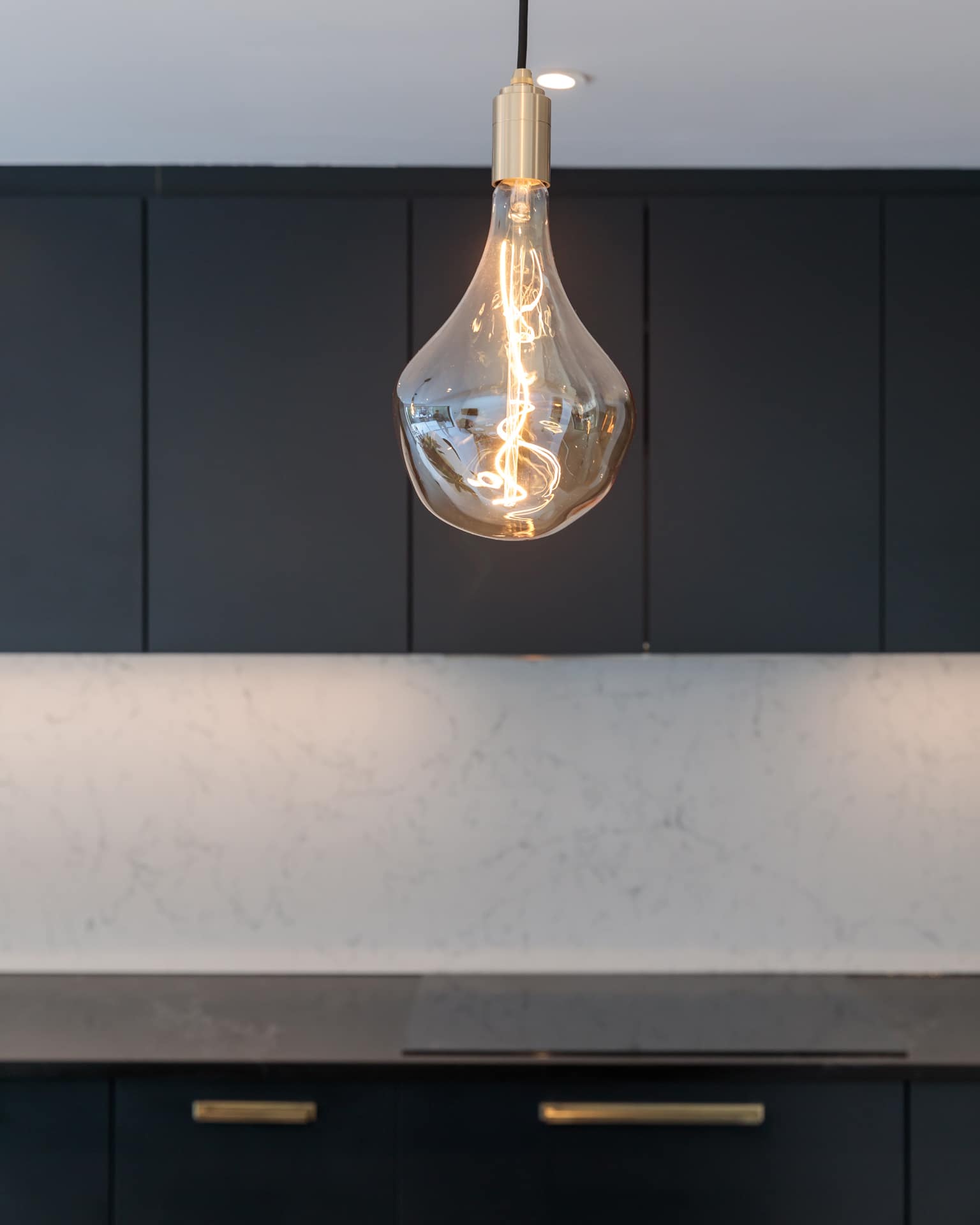 A contemporary kitchen featuring a stylish pendant light with a glowing filament, set against dark cabinetry and a light marble countertop, showcasing modern interior design elements.
