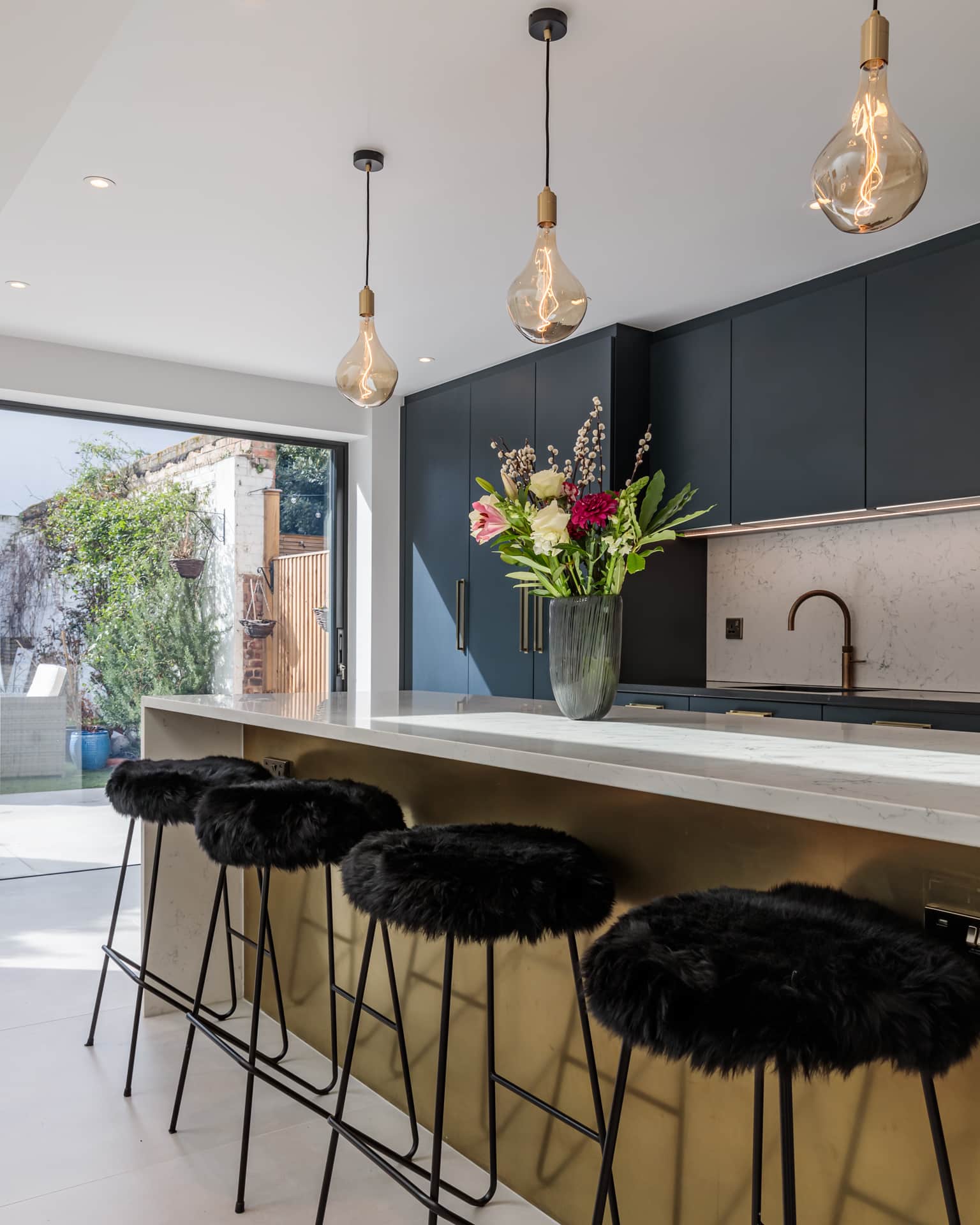 Modern kitchen featuring a sleek design with navy blue cabinetry, a marble countertop, and stylish pendant lighting. A vase of fresh flowers adds a pop of colour to the space. The kitchen island is complemented by plush black bar stools, creating an inviting atmosphere. An Arch KBB team member is present, showcasing the kitchen's functionality. Natural light streams in through large sliding doors, enhancing the contemporary feel.