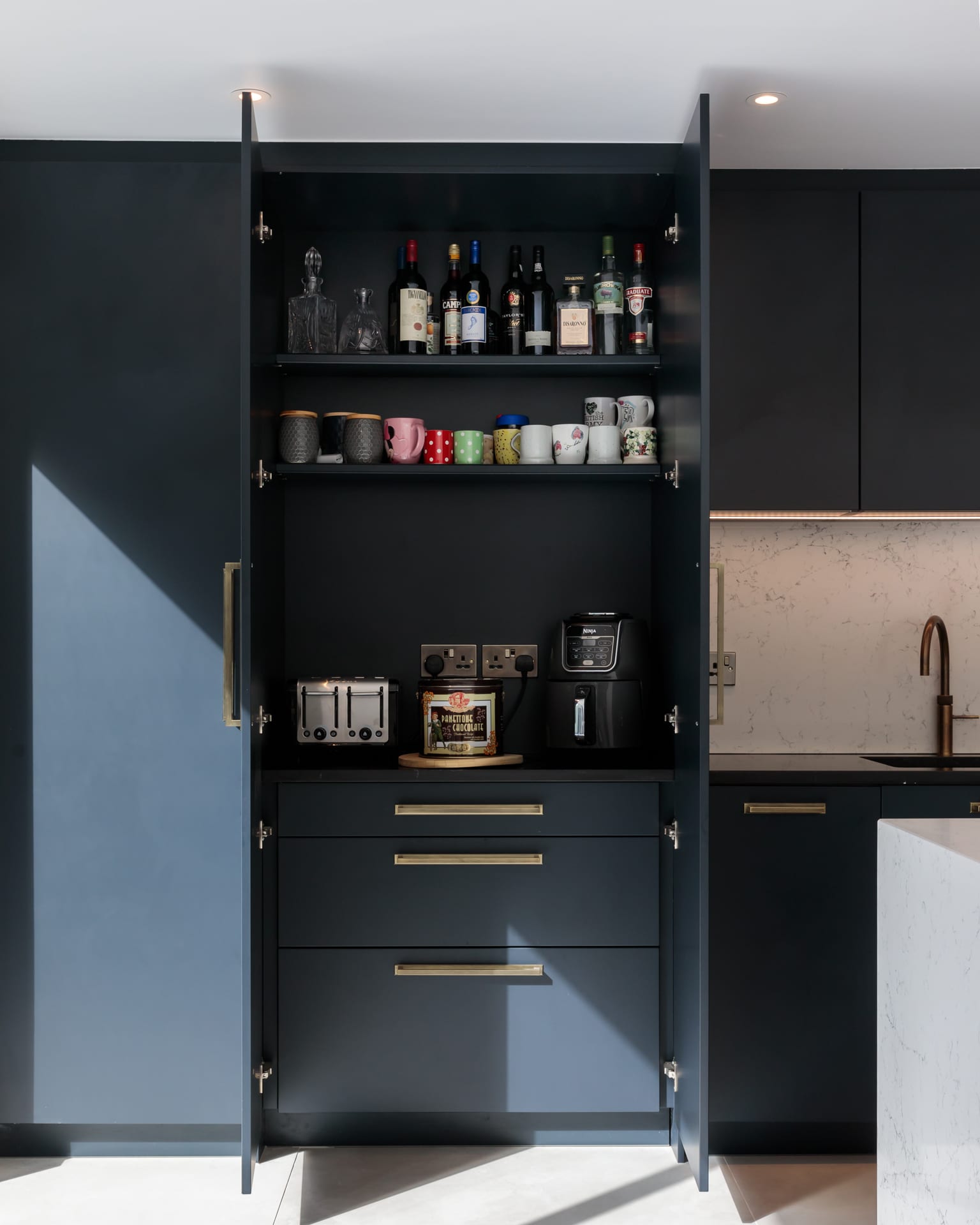 Modern kitchen interior featuring dark cabinetry, glassware, and a selection of beverages on display. An Arch KBB team member is present, showcasing the stylish and functional design elements, including a toaster and coffee machine, alongside a sleek countertop and sink.