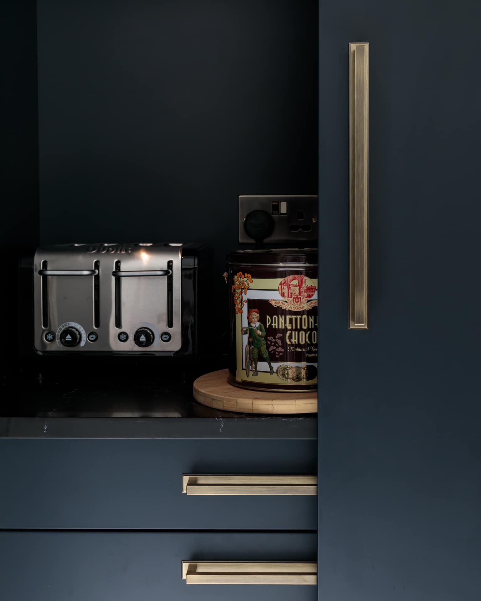 A sleek kitchen cabinet featuring a stainless steel toaster and a decorative tin of chocolate, with a wooden serving board underneath, showcasing modern design elements.