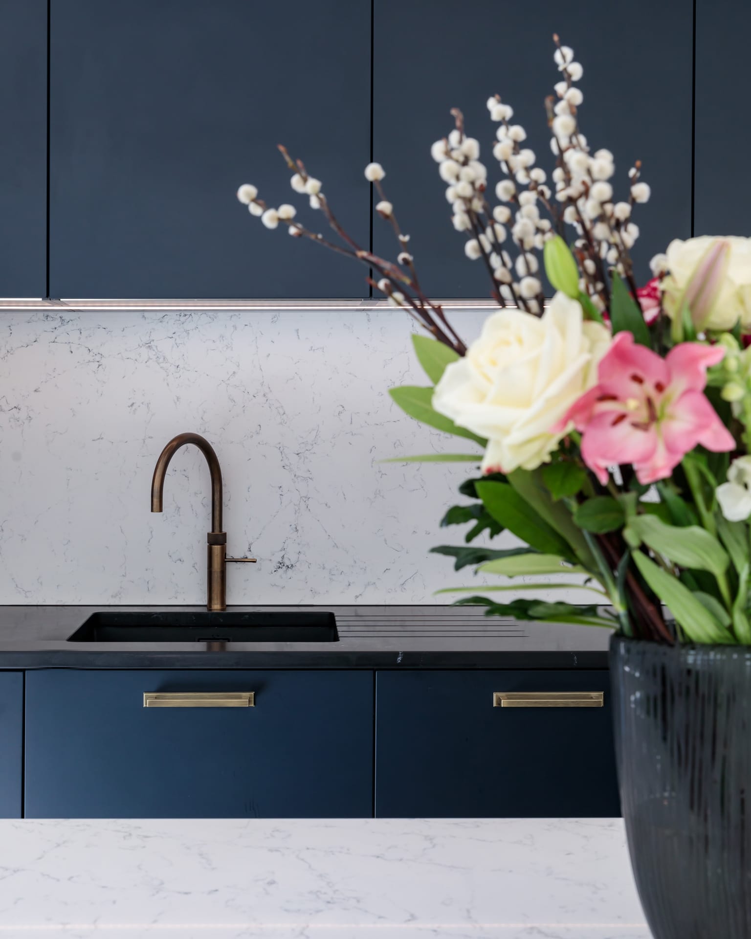 Modern kitchen featuring a sleek black sink and stylish bronze tap, with a backdrop of elegant white marble and navy cabinetry. A vibrant floral arrangement adds a touch of colour and freshness to the contemporary design. Arch KBB team member visible in the scene.