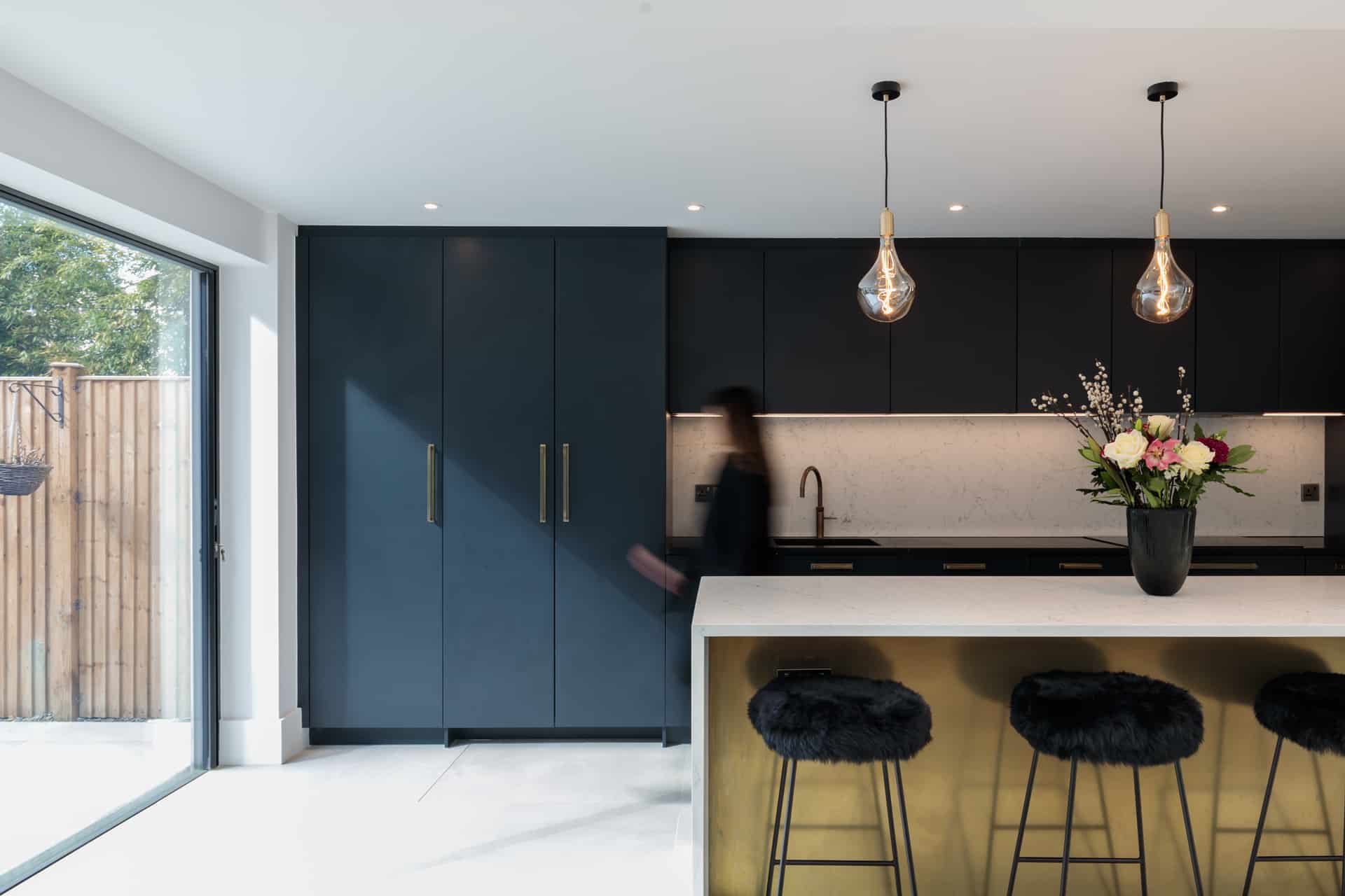 Modern kitchen design featuring sleek navy cabinetry and stylish gold accents. An Arch KBB team member is present, enhancing the contemporary atmosphere. The space is illuminated by pendant lighting, showcasing a minimalist aesthetic with a focus on functionality and elegance.