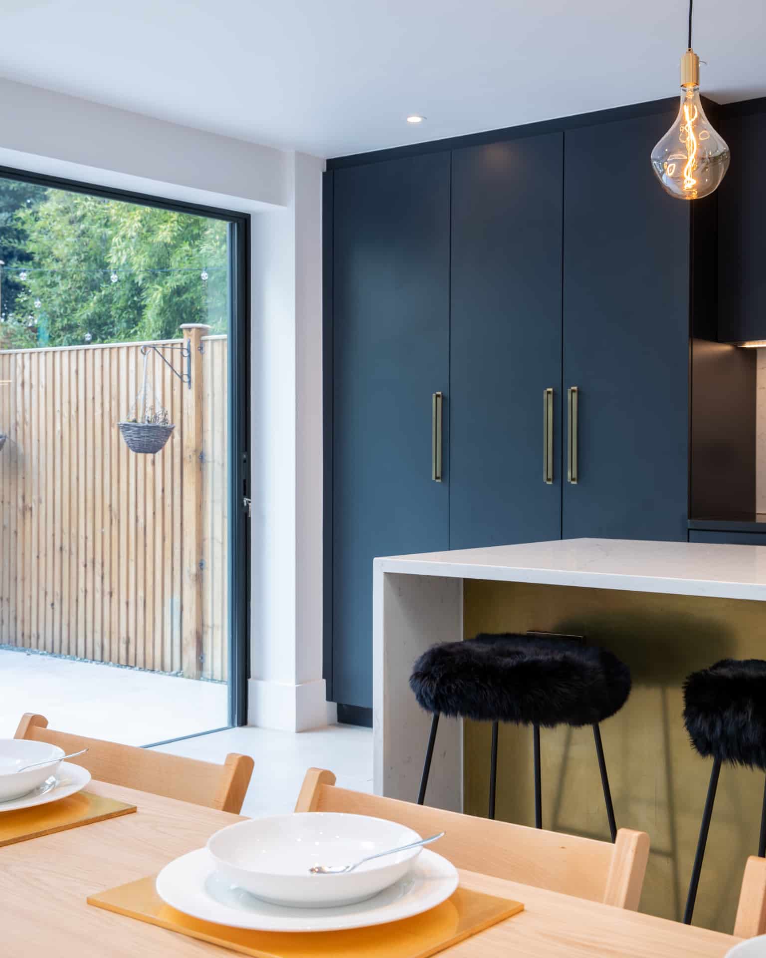 Modern kitchen featuring sleek navy cabinetry with gold handles, a stylish white countertop, and plush black bar stools. A large glass door opens to a wooden fence outside, creating a bright and airy atmosphere. An Arch KBB team member is present, showcasing the contemporary design.
