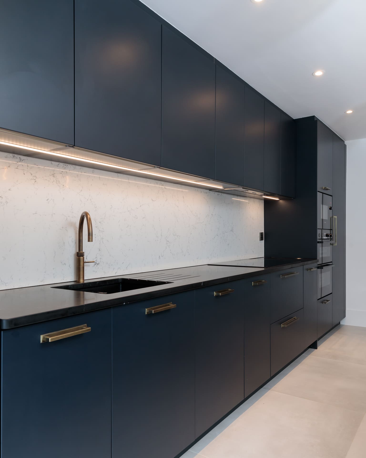 Modern kitchen design featuring sleek navy cabinetry, a minimalist black countertop, and a stylish brass tap. The space is illuminated by under-cabinet lighting, highlighting the elegant marble splashback. An Arch KBB team member is present, showcasing the kitchen's contemporary aesthetic.