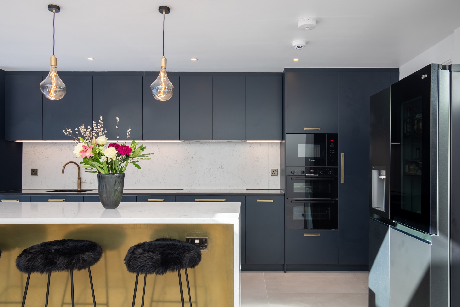 Modern kitchen design featuring dark cabinetry, marble backsplash, and gold accents. A stylish island with a white countertop and black furry stools adds elegance. Fresh flowers in a vase enhance the decor. An Arch KBB team member is present, showcasing the kitchen's functionality and aesthetic appeal.