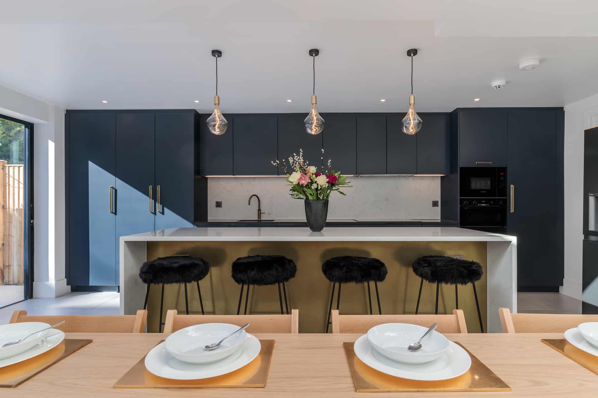 Modern kitchen featuring a sleek design with dark cabinetry, a marble countertop, and stylish bar stools. A vase of flowers adds a touch of elegance, while pendant lights illuminate the space. An Arch KBB team member is present, enhancing the contemporary atmosphere.