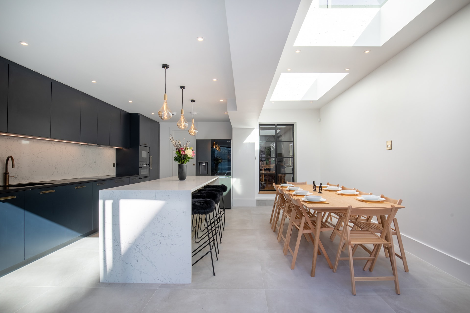 Modern kitchen interior featuring sleek black cabinetry, a marble island with bar stools, and a dining table set for a meal. Skylights allow natural light to illuminate the space, creating a bright and inviting atmosphere, with an Arch KBB team member present.