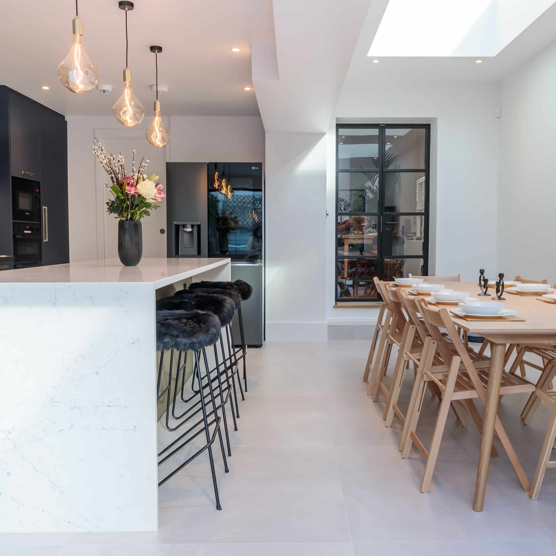 Modern kitchen and dining area featuring a marble island with bar stools, a stylish black refrigerator, and a wooden dining table set for a meal. A vase with flowers adds a touch of elegance, while large windows provide natural light. Arch KBB team member is present, enhancing the inviting atmosphere.