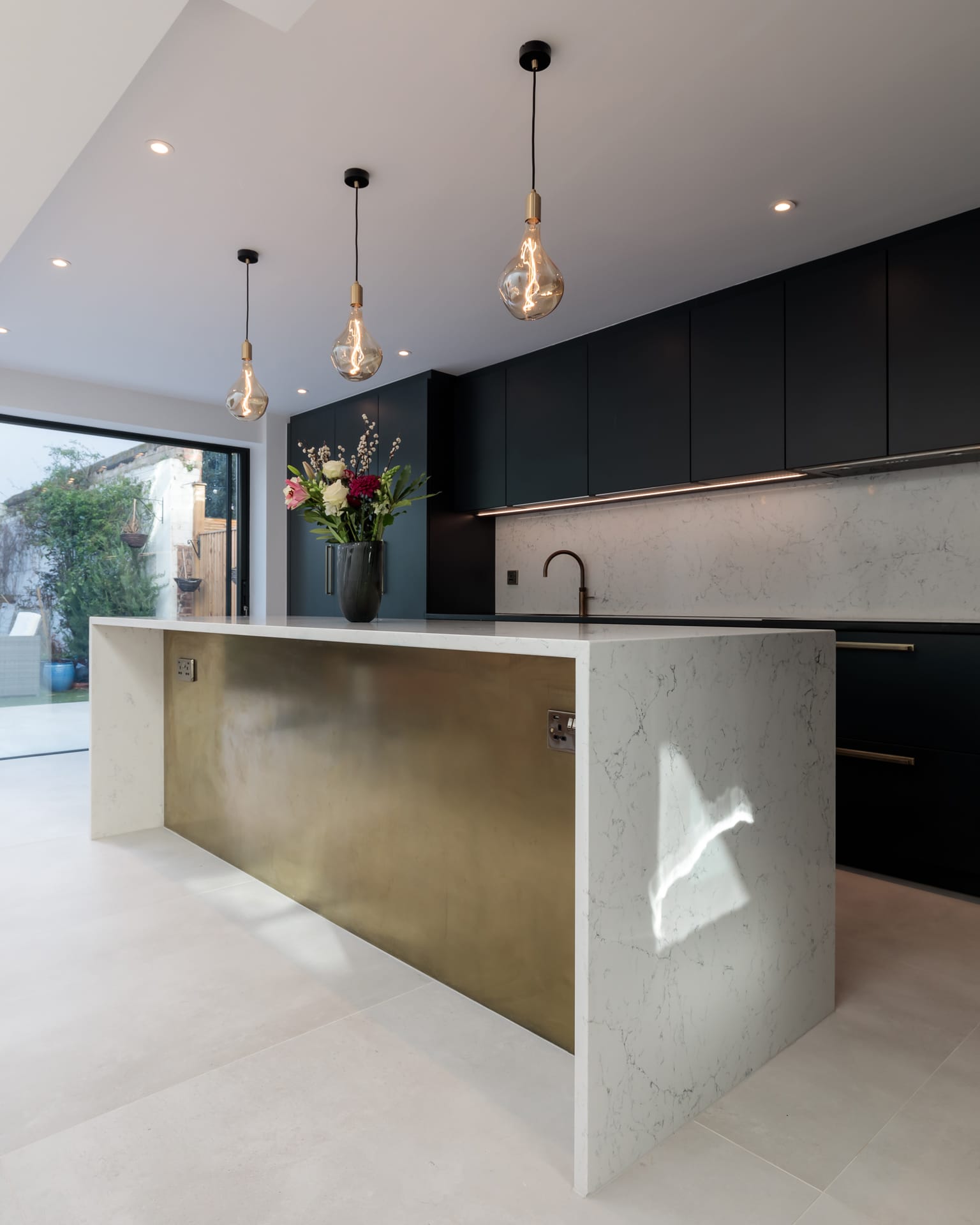 Modern kitchen featuring a stylish island with a brass front, sleek marble countertops, and dark cabinetry. Natural light floods the space through large sliding doors, highlighting a decorative vase with flowers on the island. An Arch KBB team member is present, engaged in the kitchen design.