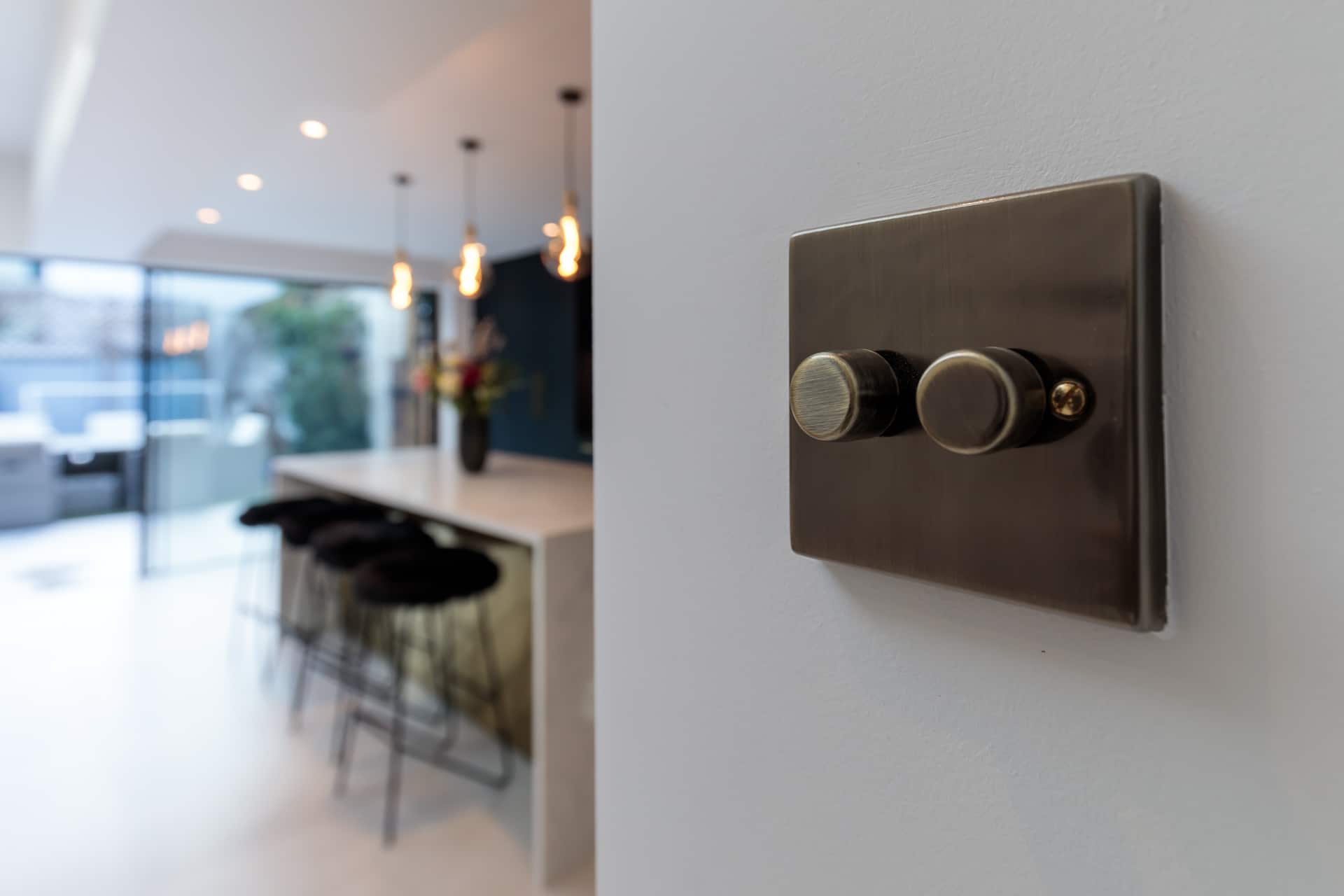 A close-up of a stylish light switch on a wall, with a modern kitchen in the background featuring a sleek countertop and bar stools. The space is well-lit and showcases contemporary design elements. An Arch KBB team member is present in the scene, contributing to the ambiance.