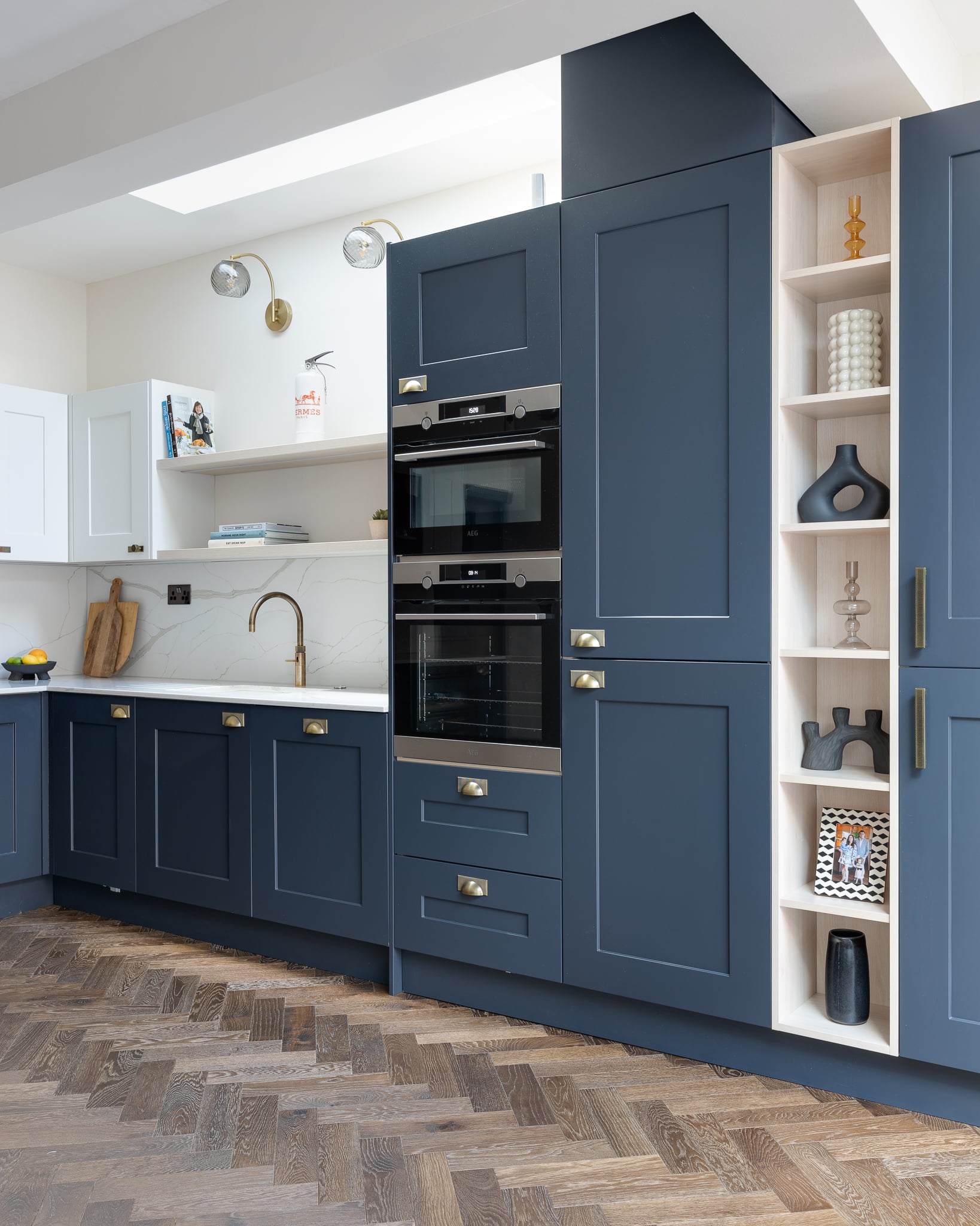 A modern kitchen featuring navy blue cabinetry, integrated double ovens, and a marble countertop. The design includes open shelving with decorative items and a stylish brass faucet. An Arch KBB team member is present, showcasing the space's functionality and aesthetic appeal.