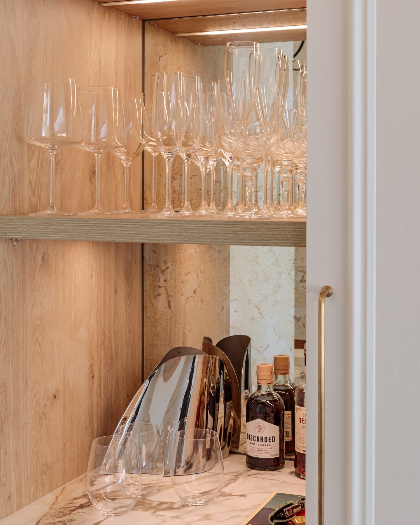 A stylish home bar featuring a collection of wine glasses neatly arranged on a wooden shelf, complemented by various bottles of spirits and a modern cocktail shaker, with warm lighting enhancing the elegant atmosphere. An Arch KBB team member is present, contributing to the inviting setting.