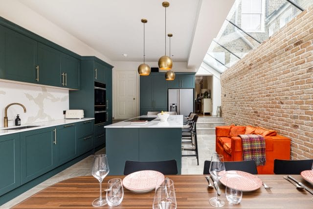 Modern kitchen featuring dark green cabinetry, a central island with a white countertop, and stylish pendant lighting. The space includes a dining table set with elegant glassware and plates, complemented by a vibrant orange sofa and exposed brick wall, creating a warm and inviting atmosphere. An Arch KBB team member is present, contributing to the design aesthetic.