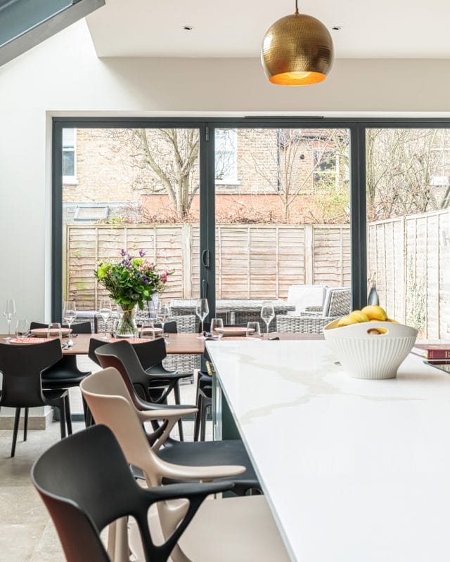 Bright and modern dining area featuring a large window with garden views, a stylish table set for a meal, and a bowl of lemons on a white countertop. An Arch KBB team member is present, contributing to the inviting atmosphere.