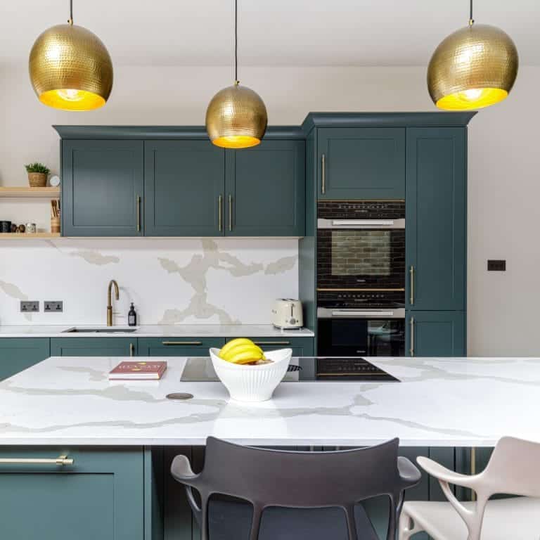 Modern kitchen interior with green cabinets, marble worktops, and brass pendant lights, featuring a breakfast bar with stylish seating and a fruit bowl.