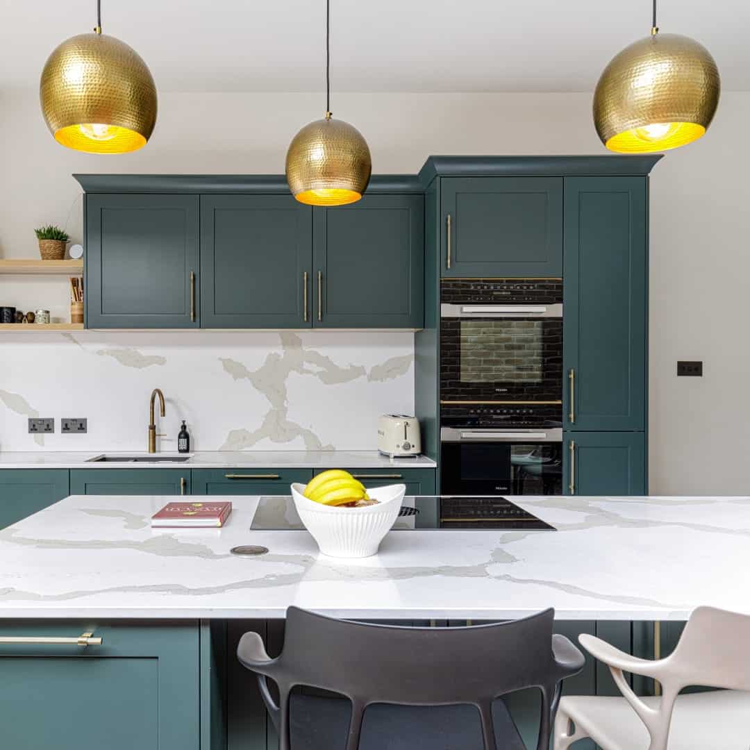Modern kitchen featuring teal cabinetry, a marble countertop, and stylish pendant lights. A bowl of fruit sits on the island, complemented by a sleek oven and kitchen appliances in the background. An Arch KBB team member is present, showcasing the design and functionality of the space.