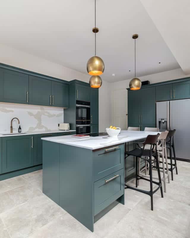 Modern kitchen featuring dark green cabinets, a large marble island, and stylish pendant lighting. The space includes built-in appliances and a seating area with bar stools. An Arch KBB team member is present, enhancing the design atmosphere.