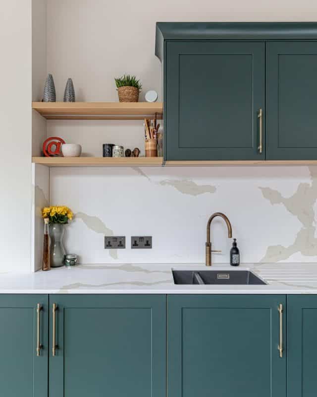 Modern kitchen featuring green cabinetry, a stylish brass tap, and a sleek sink. The countertop is adorned with a marble effect, complemented by open shelving displaying decorative items and potted plants. An Arch KBB team member is present, showcasing the design's functionality and aesthetic appeal.