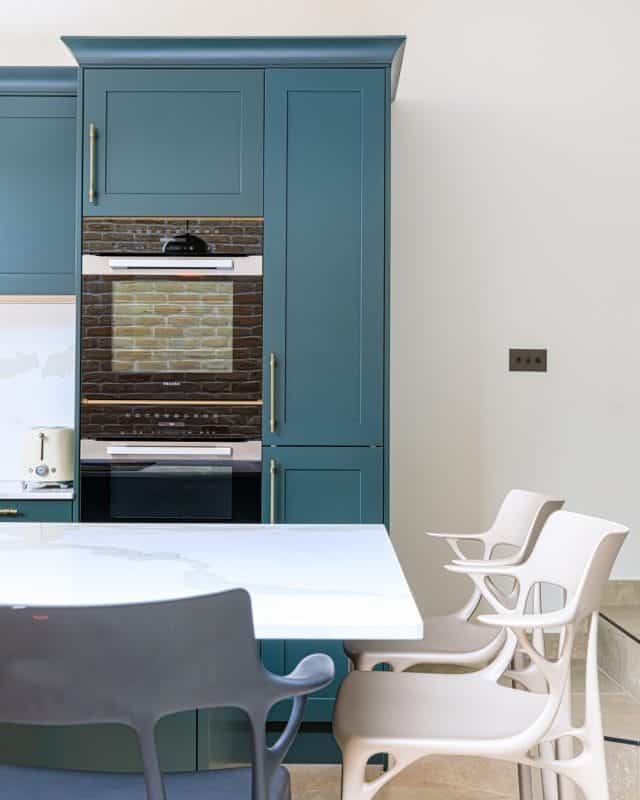 Modern kitchen featuring a sleek design with blue cabinetry, a built-in oven with a brick-style backsplash, and a minimalist white dining table. An Arch KBB team member is present, contributing to the contemporary aesthetic. The space showcases a blend of functionality and style, ideal for modern living.