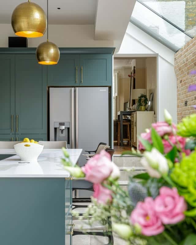Modern kitchen interior featuring a stylish island with a white bowl of lemons, green cabinetry, and a stainless steel fridge. An Arch KBB team member is visible in the background, with a dining area and plants adding warmth to the space. Bright floral arrangements enhance the inviting atmosphere.