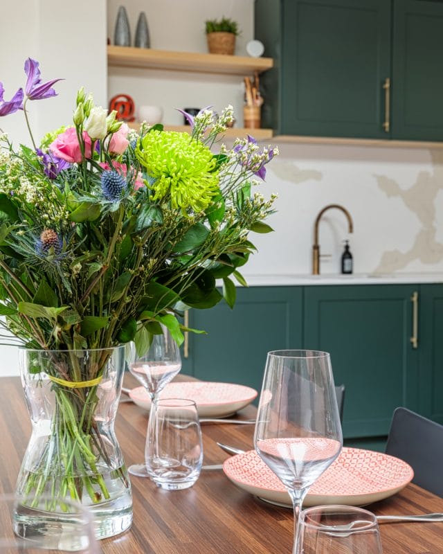 A vibrant floral arrangement in a glass vase sits on a wooden dining table, with elegant glassware and pink plates. In the background, a stylish kitchen features green cabinetry and a modern sink. An Arch KBB team member is present, enhancing the inviting atmosphere of the space.